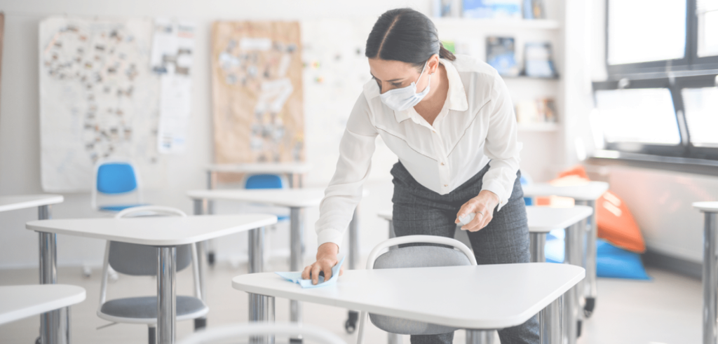 Person wearing a face mask disinfecting desks in a classroom with a cleaning cloth and spray bottle