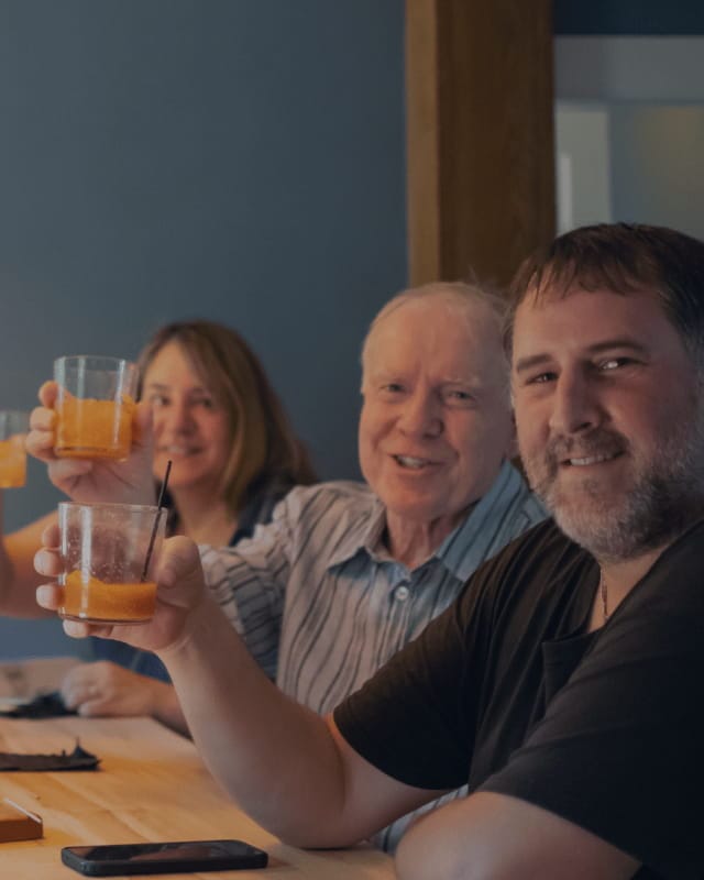 Three people sitting at a bar raising cocktail glasses and smiling at the camera in a warm, casual setting.
