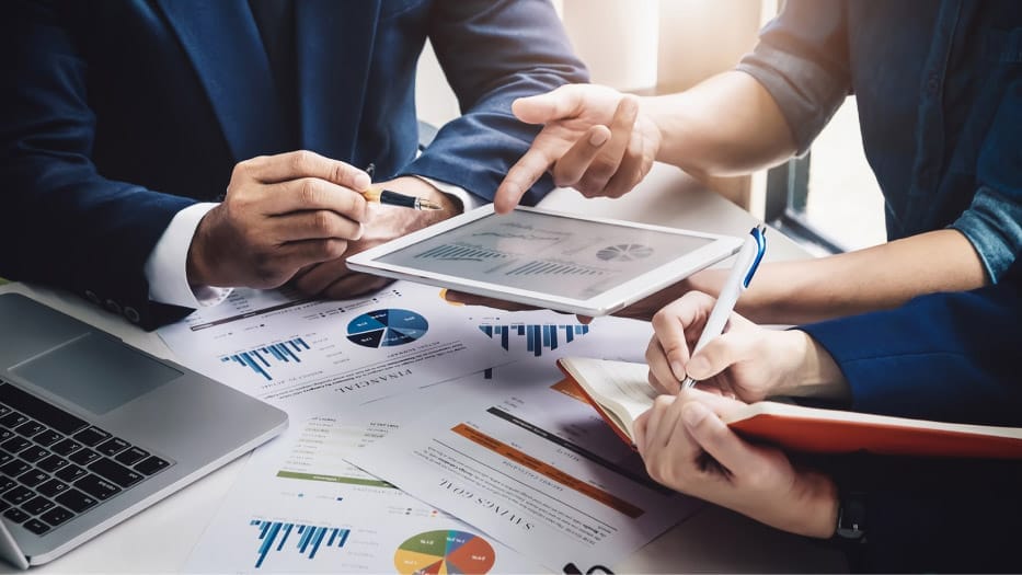 Business professionals reviewing charts and documents at a desk, with one person pointing to a tablet while another takes notes.