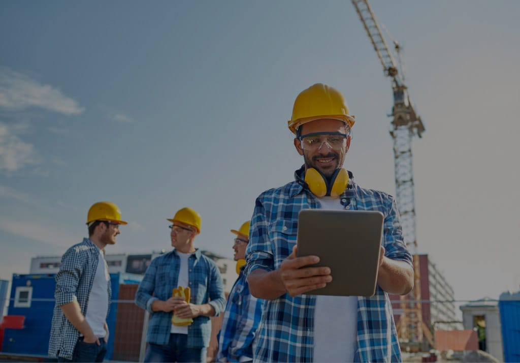 HVAC technician in a hard hat using a tablet on a job site, with crew members and construction equipment in the background.