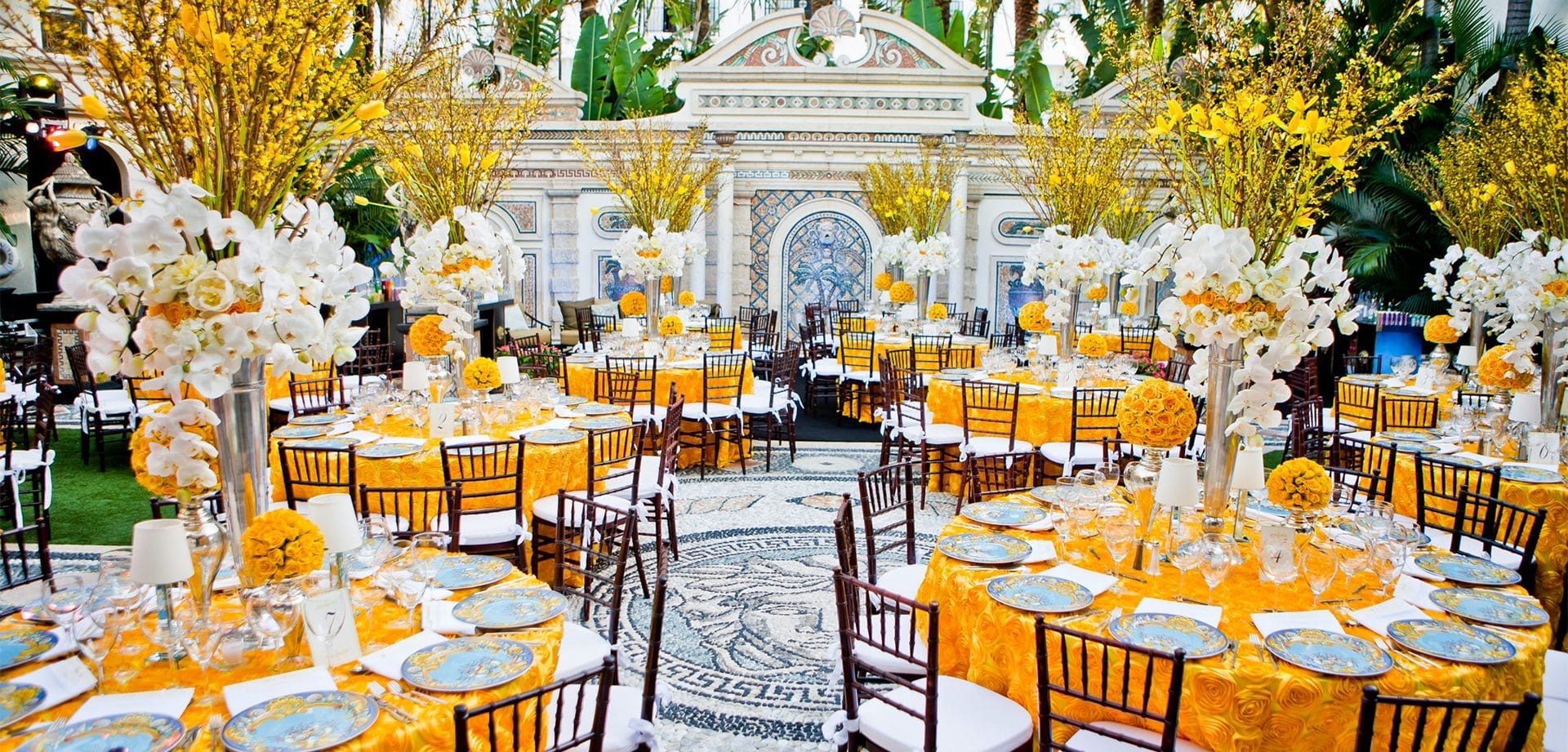 Elegant outdoor restaurant event setup with round dining tables, floral centerpieces, patterned plates, and bright yellow linens in a courtyard setting