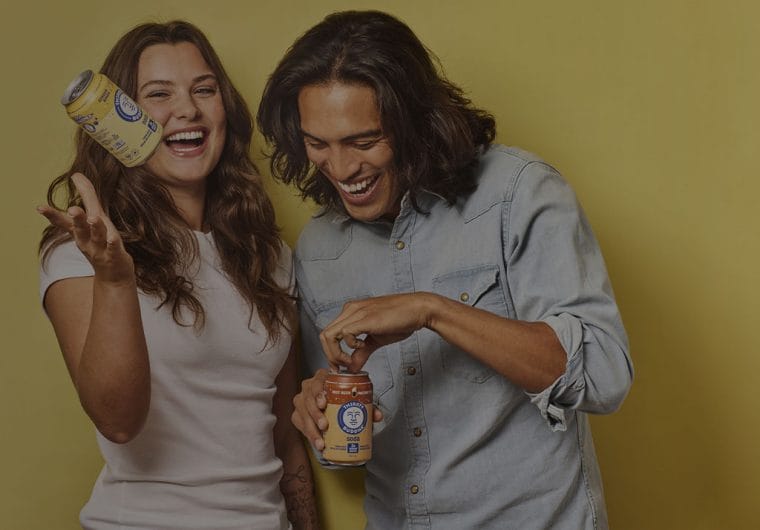 product-website-design-buddha-brands-background-image Two laughing people holding Thirsty Buddha soda cans against a warm yellow background, with one tossing a can in the air